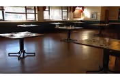 A spacious dining room with several empty tables and chairs arranged neatly. The room has large windows allowing natural light to fill the space, and the floor is made of wood. The tables are set with cups and napkins, ready for use.