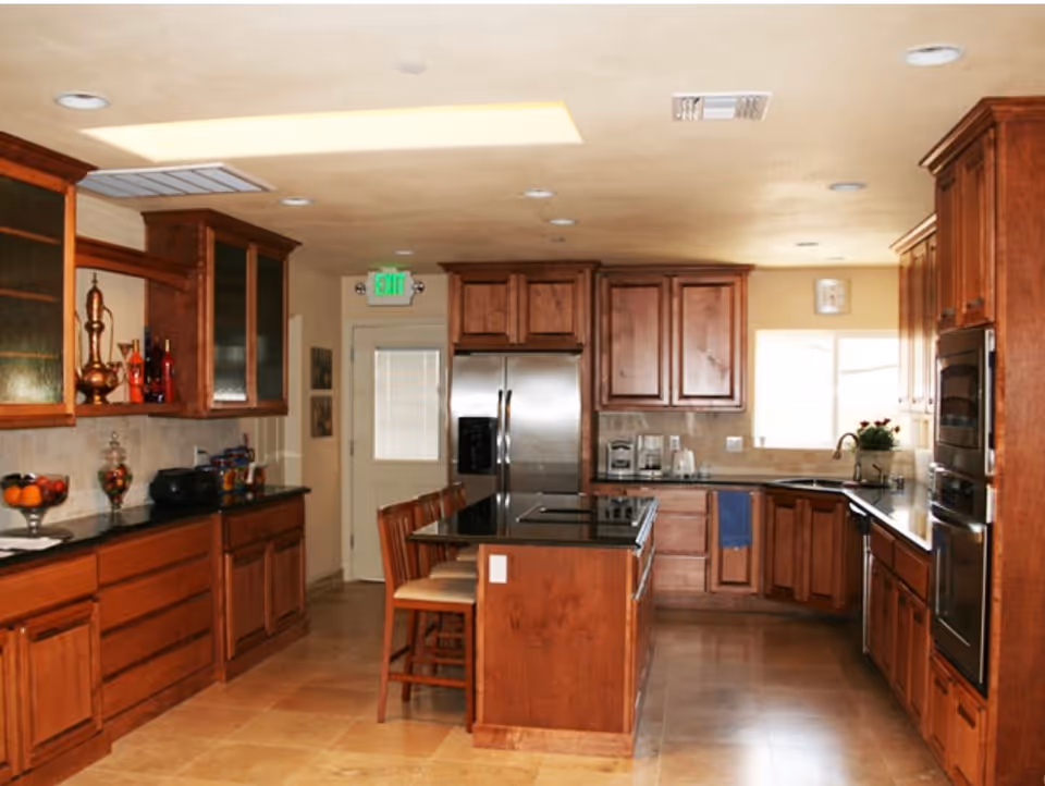 A spacious kitchen with wooden cabinets, a central island with a black countertop and two chairs, stainless steel refrigerator, built-in oven, and a window above the sink letting in natural light.