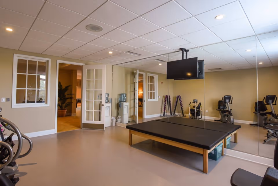 A fitness room with exercise equipment including stationary bikes, a rack of dumbbells, a padded exercise table, and a wall-mounted TV. The room has beige walls, a large mirror covering one wall, and a door leading to another area.