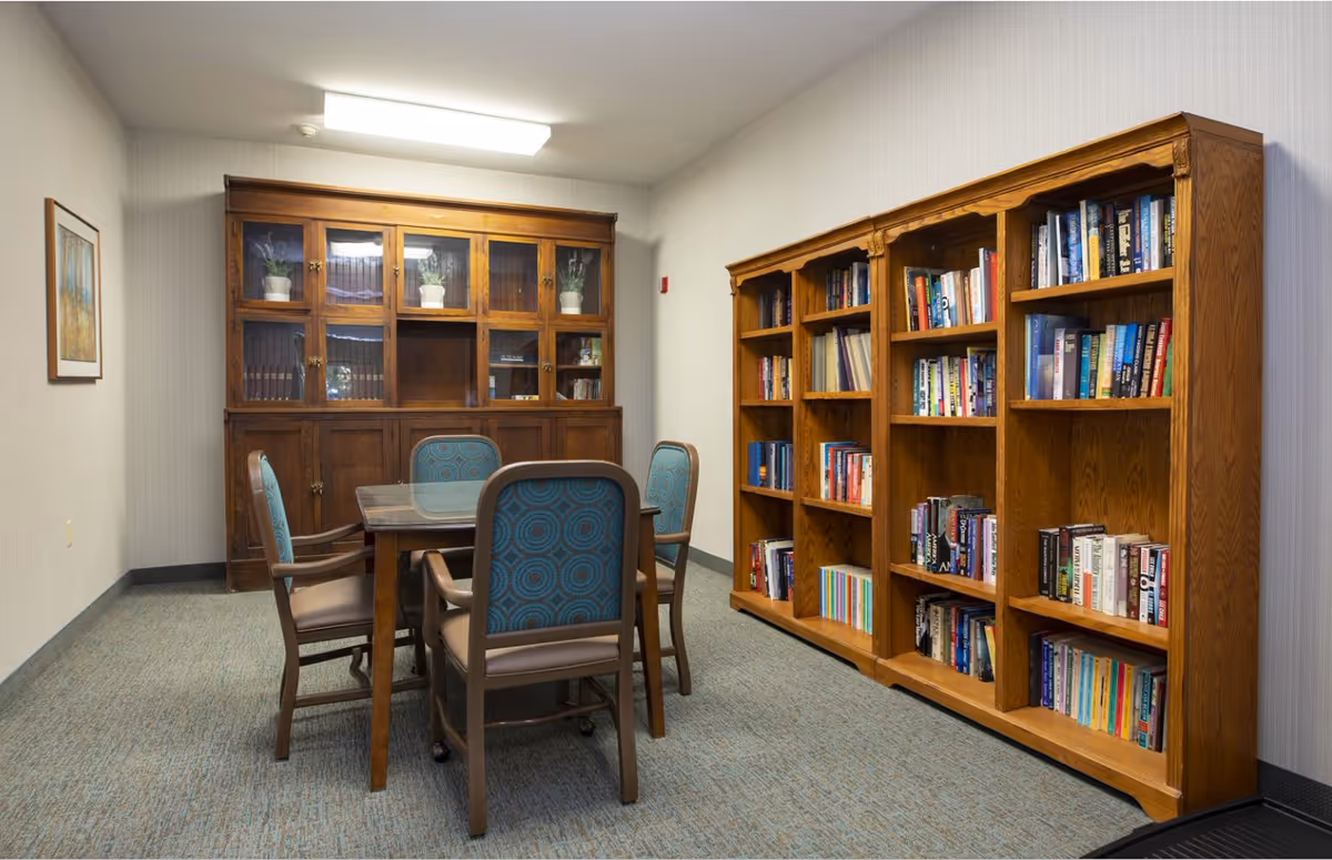 A quiet reading or meeting room with a wooden table surrounded by four chairs with blue patterned upholstery. Against one wall is a large wooden cabinet with glass doors displaying potted plants and books. On the opposite wall are three wooden bookshelves filled with various books. The room has light-colored walls and carpeted flooring.