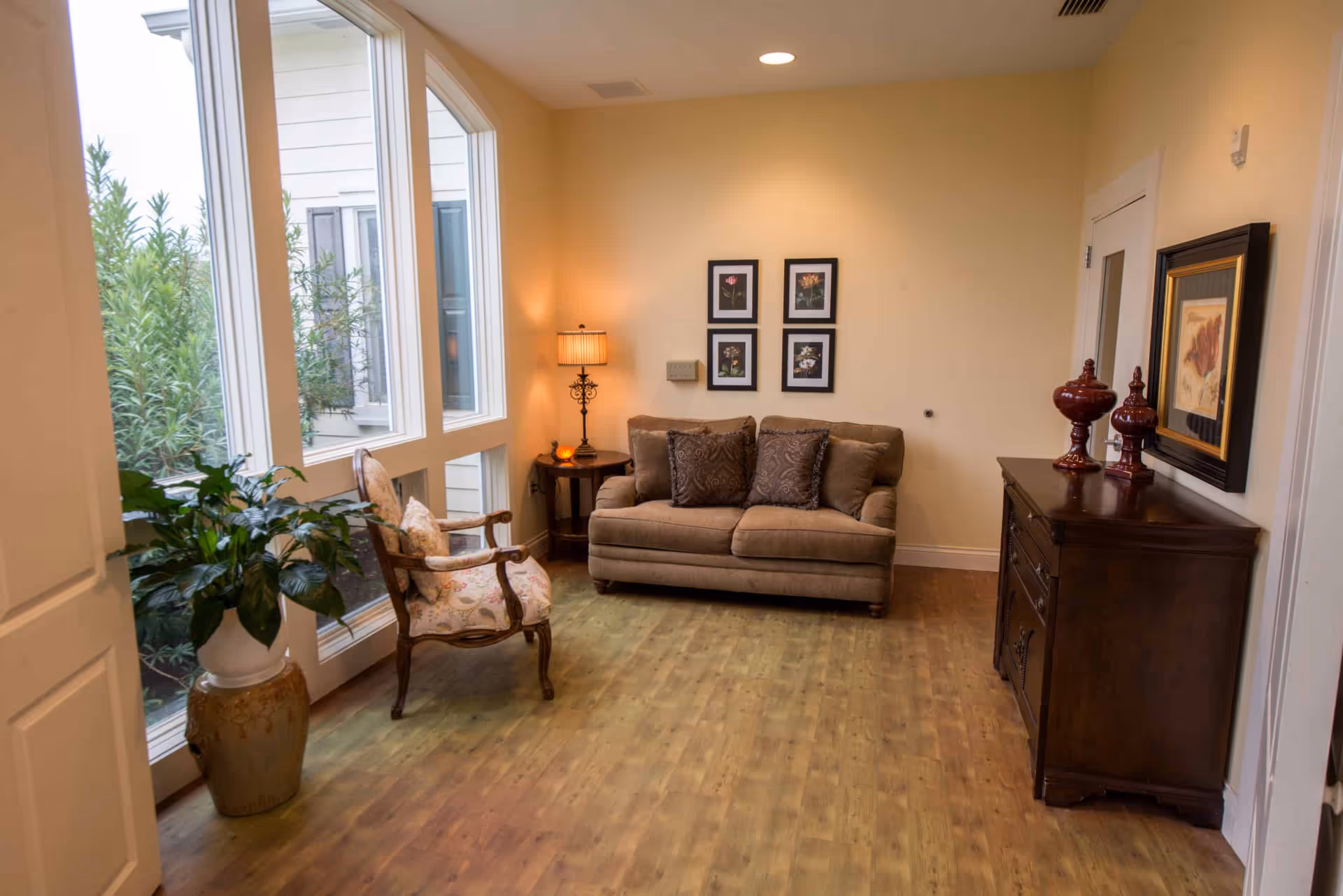 A cozy living room area with a brown sofa adorned with three decorative pillows, a wooden armchair with floral upholstery, a side table with a lamp, and a wooden cabinet with decorative items. Large windows let in natural light and show greenery outside. The walls are painted a soft yellow and decorated with framed floral artwork.