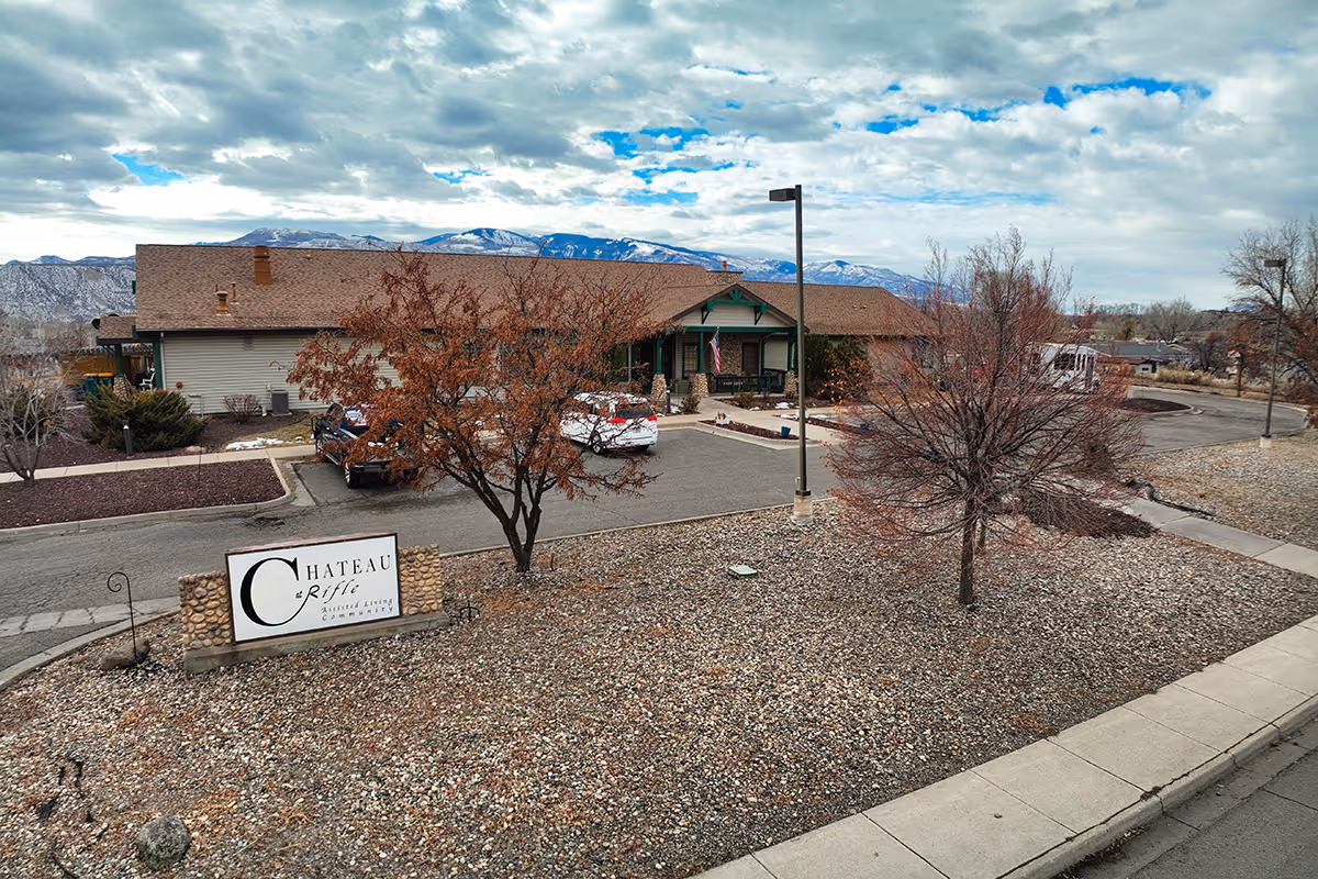 Exterior view of Chateau At Rifle assisted living community building with a parking area, leafless trees, and mountains in the background under a partly cloudy sky.