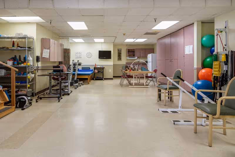 A rehabilitation therapy room with exercise equipment, therapy tables, chairs, colorful exercise balls, and various physical therapy tools organized on shelves and racks. The room has a beige floor, fluorescent ceiling lights, and cabinets along the walls.