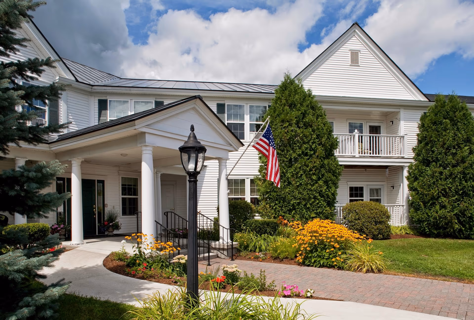 Exterior view of a white two-story building with a covered entrance supported by columns, an American flag, well-maintained garden with colorful flowers, and a black lamp post in front under a partly cloudy sky.