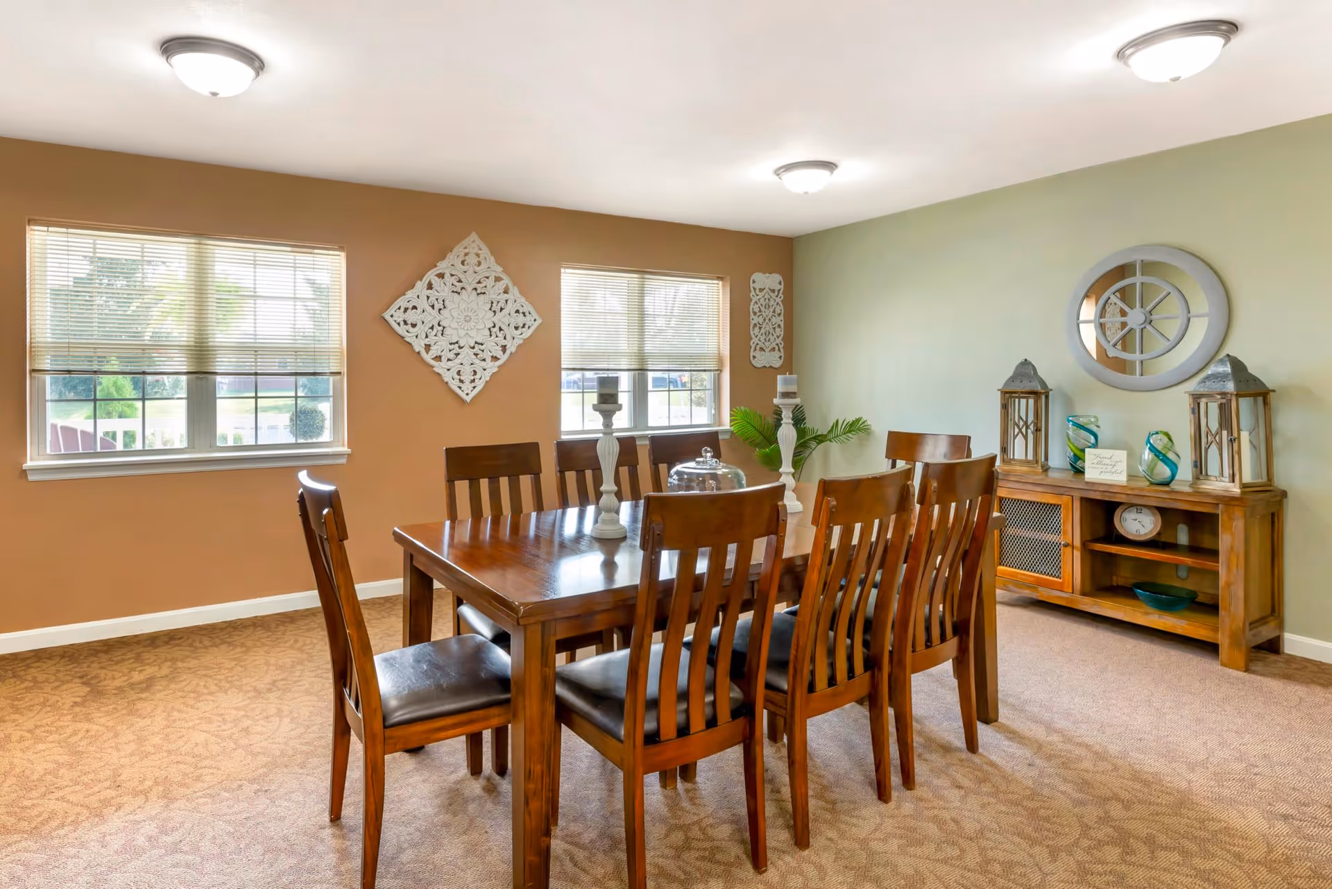Well-lit dining room with a wooden table surrounded by chairs, a sideboard, windows, and decorative wall accents.