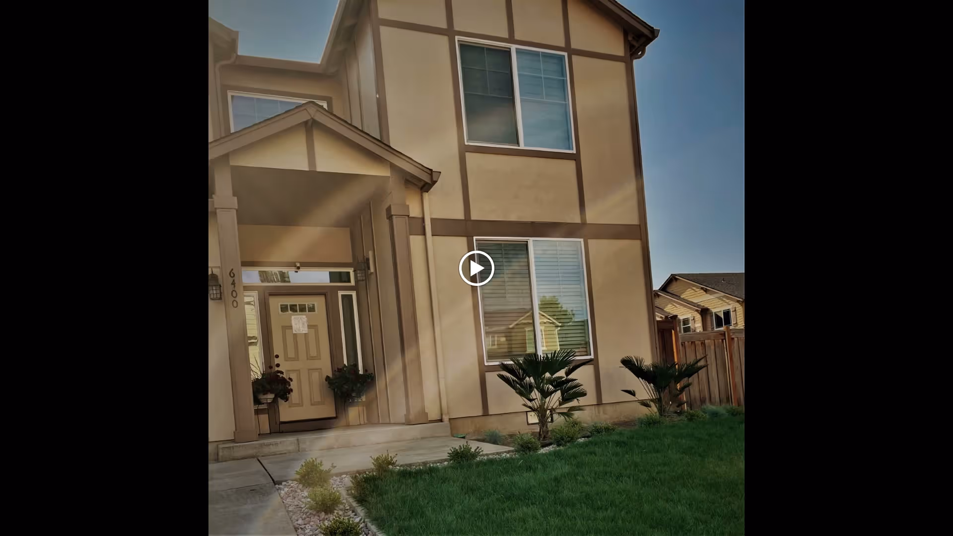 Front exterior view of a two-story beige house with brown trim, a small covered porch with two potted plants, a green lawn, and a wooden fence on the right side.