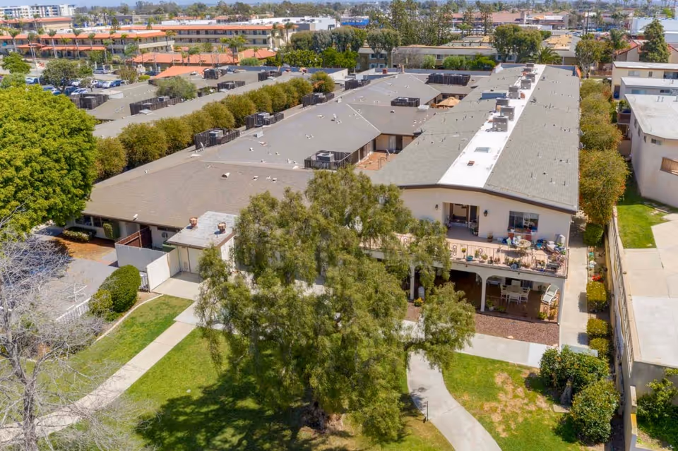 Aerial view of a large assisted living facility building with a gray roof, surrounded by trees and green lawns. The building has multiple units with balconies and patios, and there are paved walkways around the property. Other buildings and parking areas are visible in the background under a clear sky.