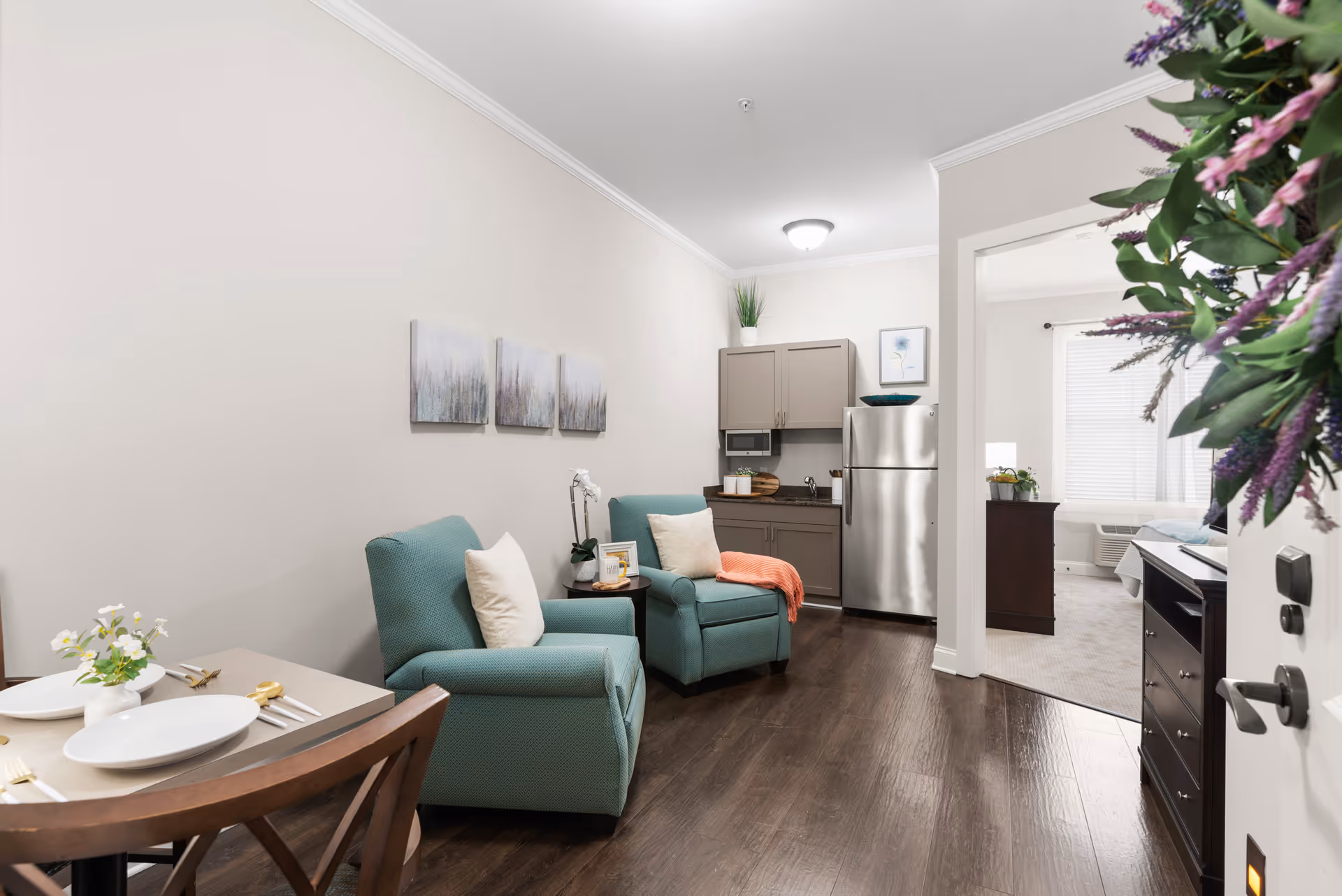Studio-style living room with two teal armchairs, a small dining table, a kitchenette with stainless steel refrigerator, and a view into the bedroom.