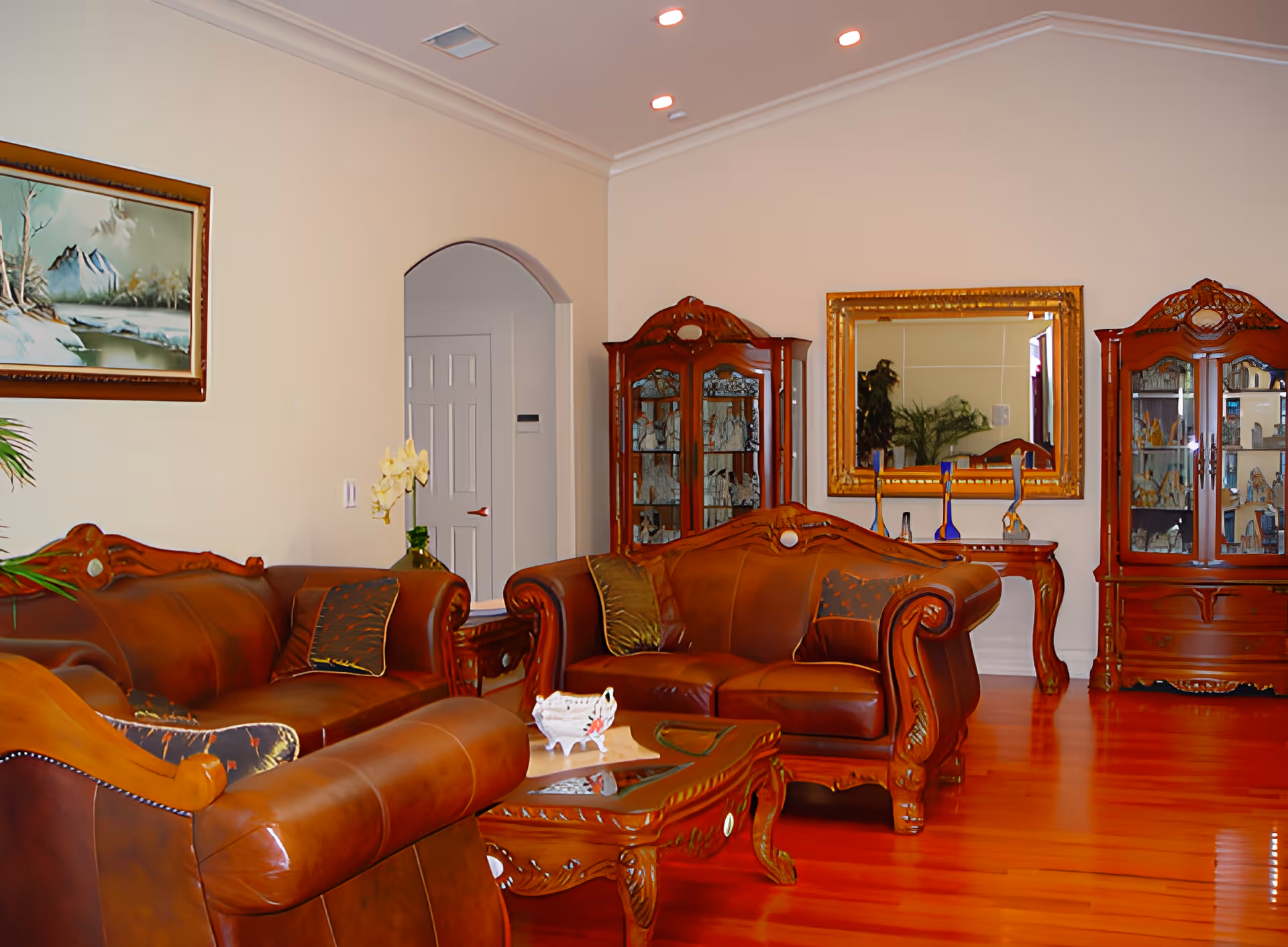 A formal living room with brown leather sofas, ornate wooden coffee table, glass-front display cabinets and a large framed mirror on hardwood floors.