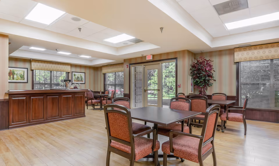 Bright communal dining area with several tables and upholstered chairs, a wooden service counter, and glass doors leading outside.