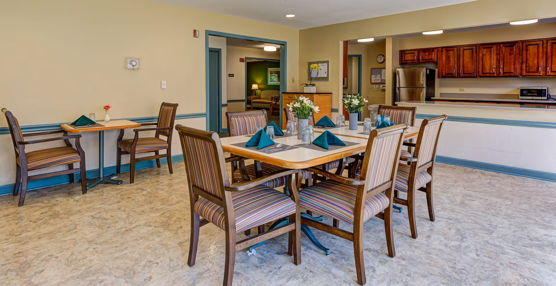 A dining area in a senior living facility with a square table set for six people. The table has striped cushioned chairs, teal folded napkins, glasses, and small flower arrangements. In the background, there is a kitchen area with wooden cabinets, a stainless steel refrigerator, and a microwave. Additional seating with two small tables and chairs is visible along the wall.