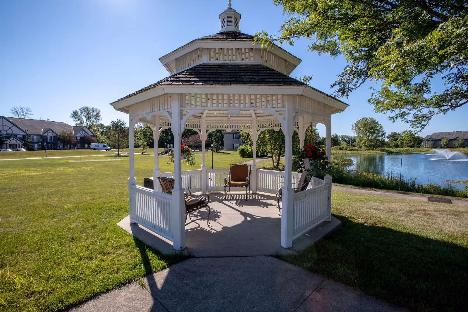 A white wooden gazebo with chairs inside situated on a concrete pad in a grassy area near a pond with a fountain. Trees and residential buildings are visible in the background under a clear blue sky.