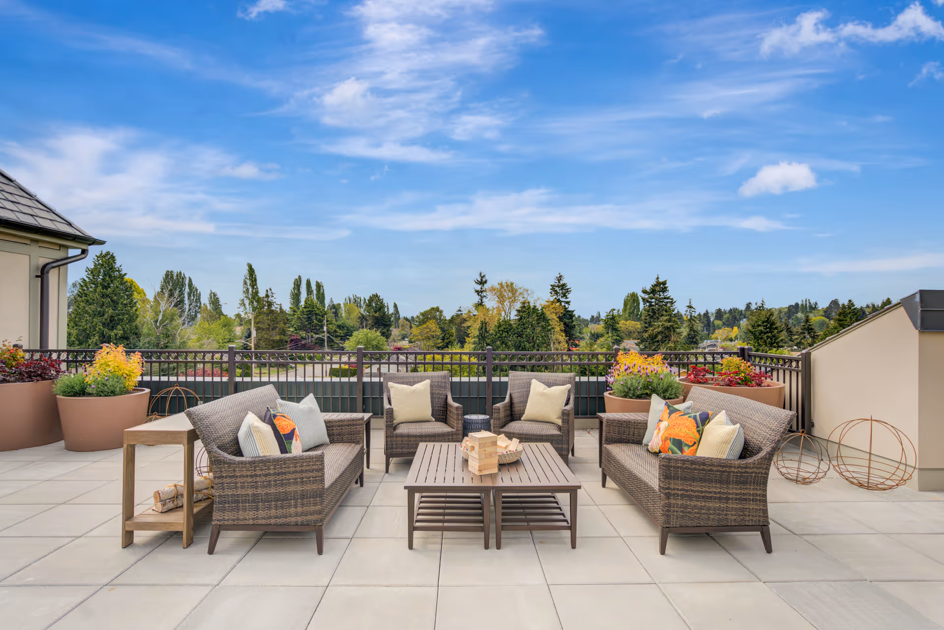 Outdoor patio area with wicker sofas and chairs arranged around a wooden coffee table. The seating has colorful cushions, and large planters with flowers are placed along the railing. The background shows a clear blue sky and green trees.