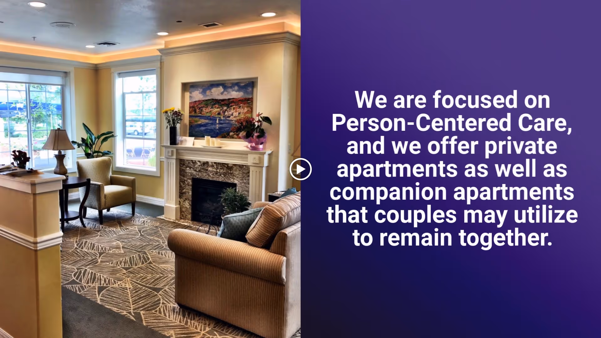 A cozy living room area with beige walls, a patterned carpet, a fireplace with a painting above it, a sofa, armchairs, a table lamp, and plants near large windows letting in natural light.