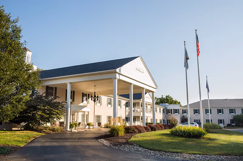 Exterior view of a senior living facility building with a large covered entrance supported by white columns, surrounded by well-maintained landscaping including bushes and grass. Three flagpoles with flags are visible on the right side under a clear blue sky.