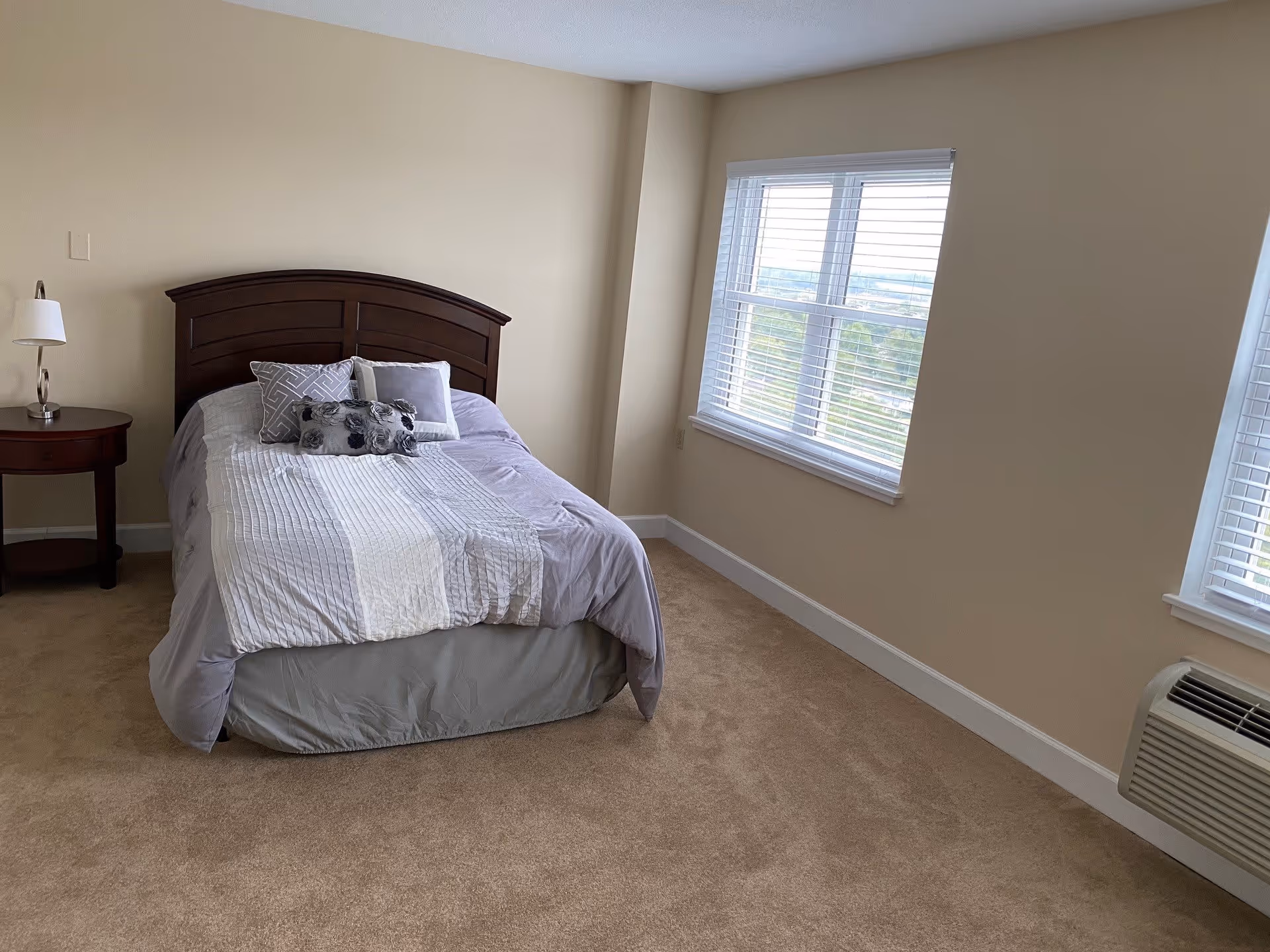 A bedroom with a bed that has a wooden headboard and is made with a gray and white striped comforter and decorative pillows. There is a small round wooden nightstand with a lamp on the left side of the bed. The room has beige walls, carpeted floor, and two windows with white blinds letting in natural light.