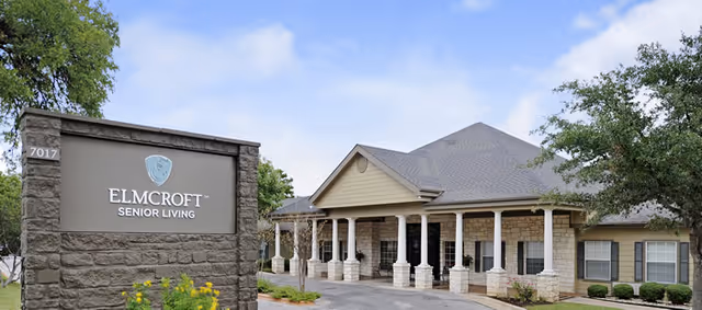 Exterior view of Elmcroft Senior Living facility showing a stone sign with the facility name and logo, a driveway, and a building with a covered entrance supported by white columns under a partly cloudy sky.