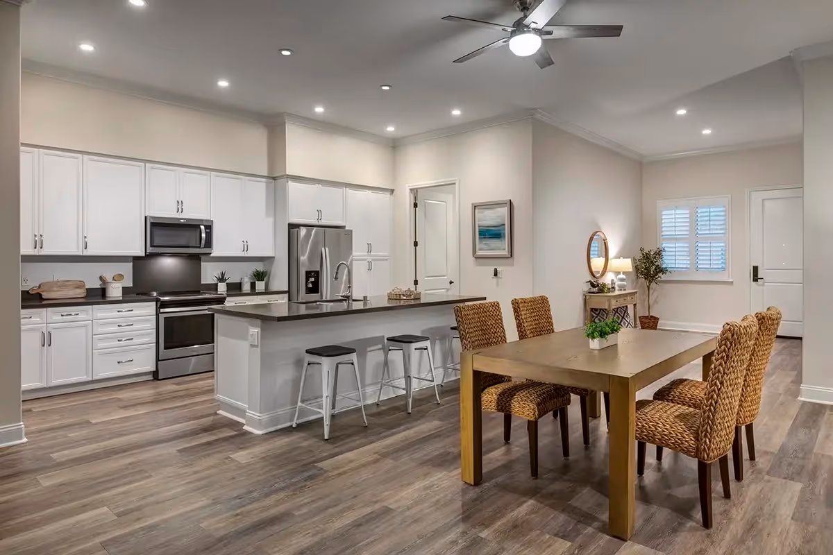 Open-concept kitchen and dining area with white cabinets, stainless steel appliances, an island with stools, and a wooden table with woven chairs.