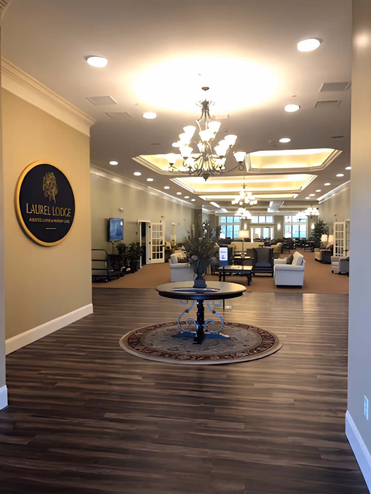 Interior view of Laurel Lodge Assisted Living & Memory Care featuring a spacious common area with wooden flooring, a round table with a floral arrangement on a circular rug in the foreground, multiple chandeliers hanging from the ceiling, and comfortable seating areas with sofas and chairs in the background.