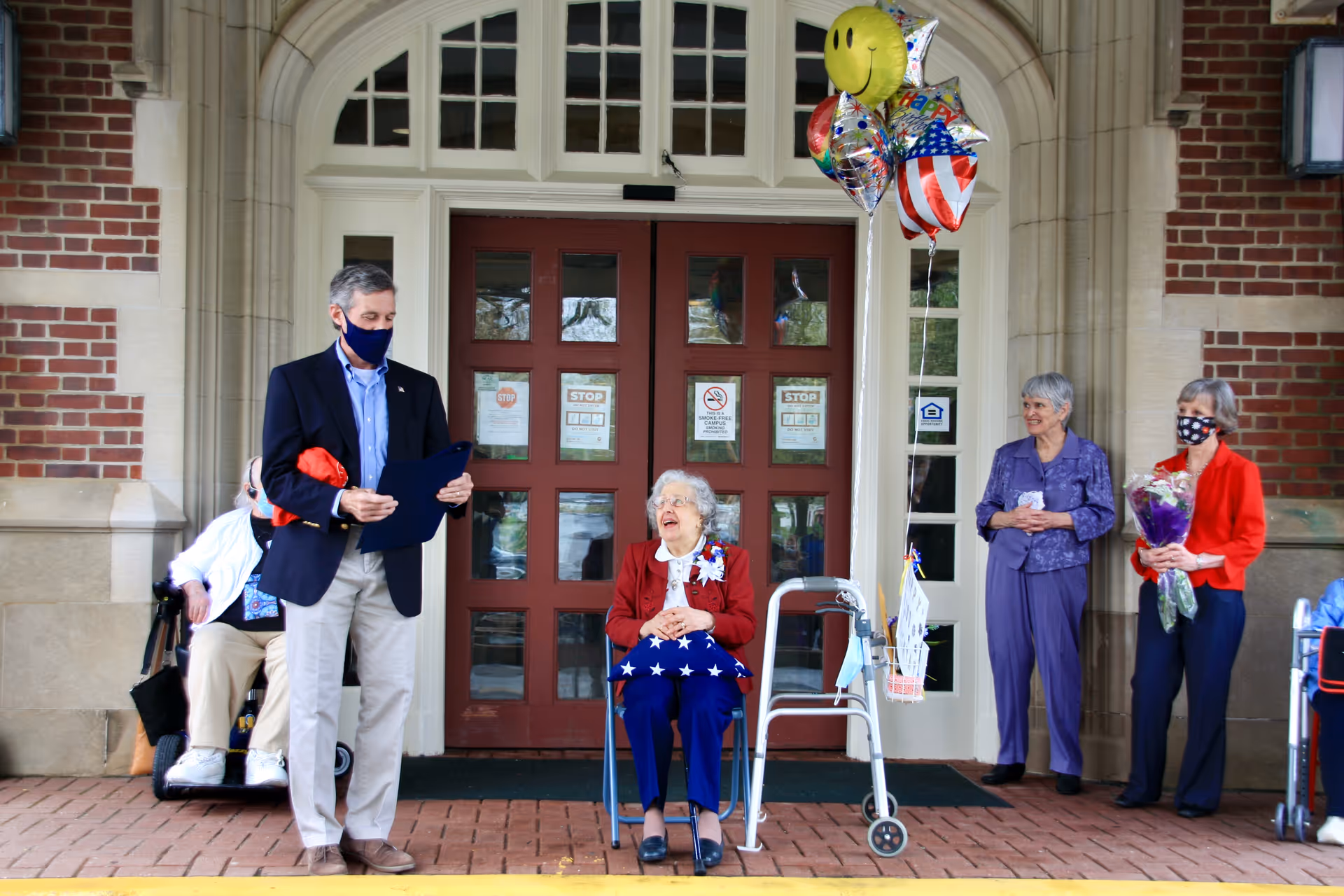 An elderly woman seated in a chair outside a building entrance, holding a folded American flag, surrounded by three standing adults, one holding balloons and another holding a bouquet of flowers. One man is reading from a document. The entrance has double red doors with glass panels and signs posted on them.