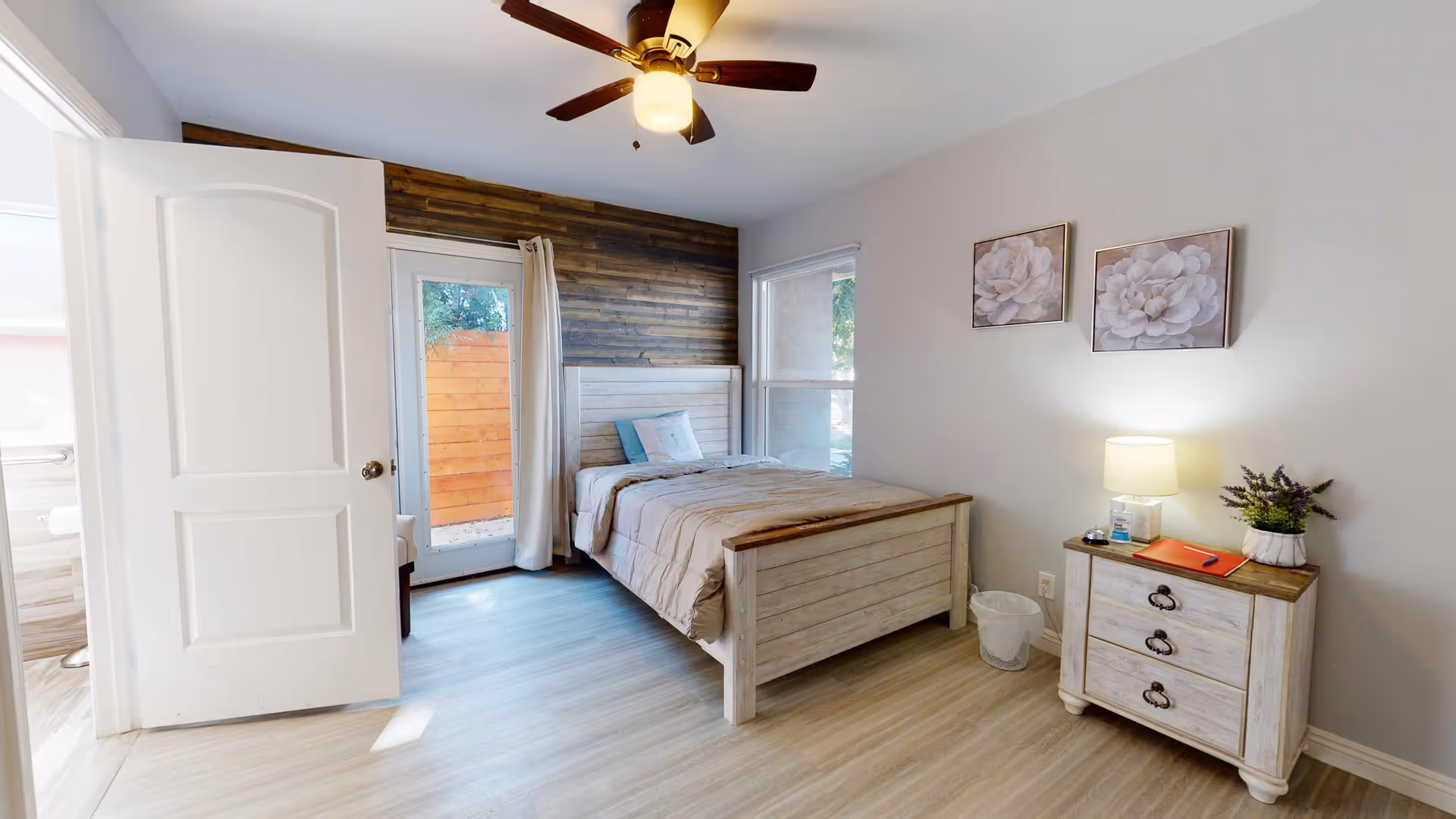 A bright bedroom featuring a single bed with beige bedding and blue pillows, a wooden nightstand with a lamp, plant, and some items on it, two floral paintings on the wall, a ceiling fan with light, a window, and a glass door leading outside with a curtain. The floor is light wood, and the walls are painted light gray. An open door leads to a bathroom.