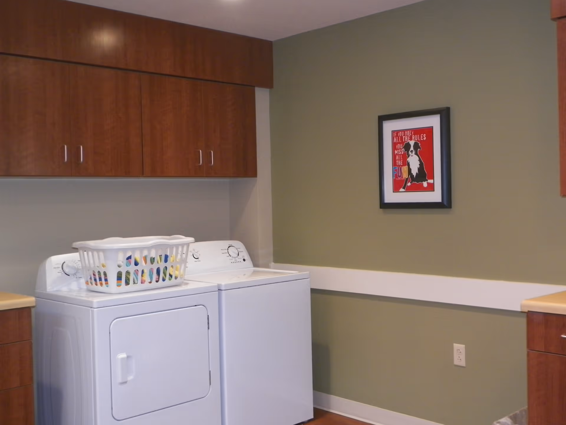Laundry room with a white washing machine and dryer side by side, a laundry basket on top of the dryer, wooden cabinets above the machines, and a framed picture of a dog on a green wall.