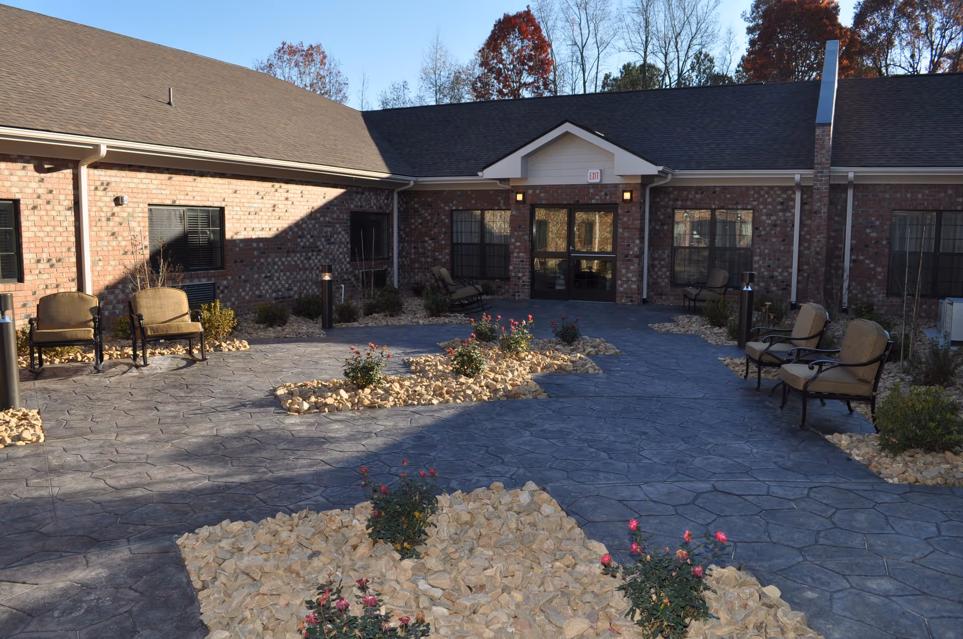 Brick single-story senior living facility courtyard with stone-paved walkways, rock landscaping with flowering shrubs and outdoor seating by the entrance.