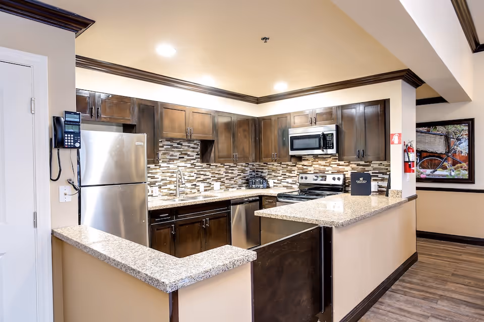 Modern kitchen with dark wooden cabinets, stainless steel refrigerator, dishwasher, stove, and microwave. The kitchen features a granite countertop with a breakfast bar, a mosaic tile backsplash, and recessed ceiling lights. A fire extinguisher is mounted on the wall near a framed picture of a bicycle with a basket.