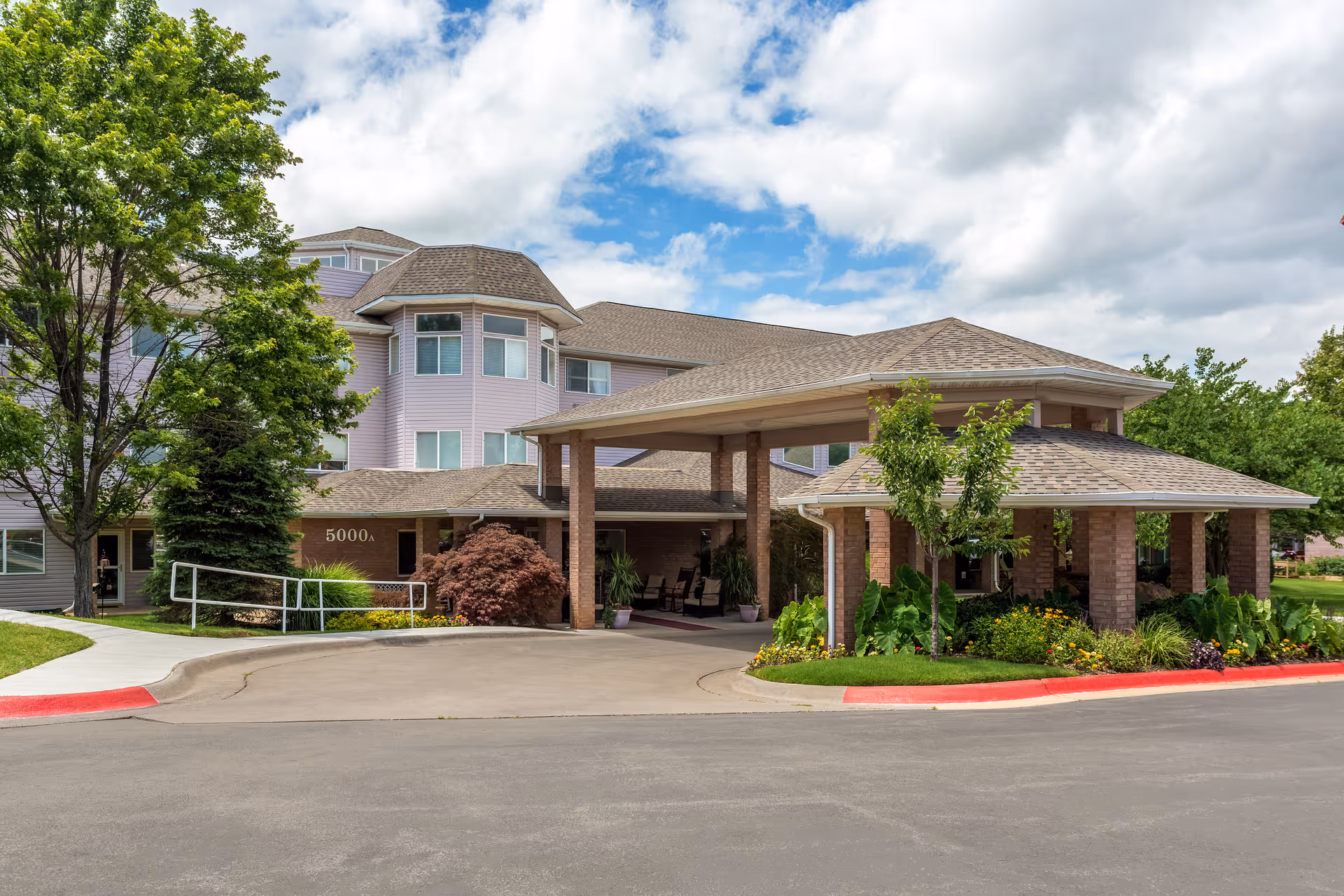 Exterior view of a senior living facility building with a covered entrance supported by brick columns, surrounded by trees, shrubs, and a well-maintained garden under a partly cloudy sky.