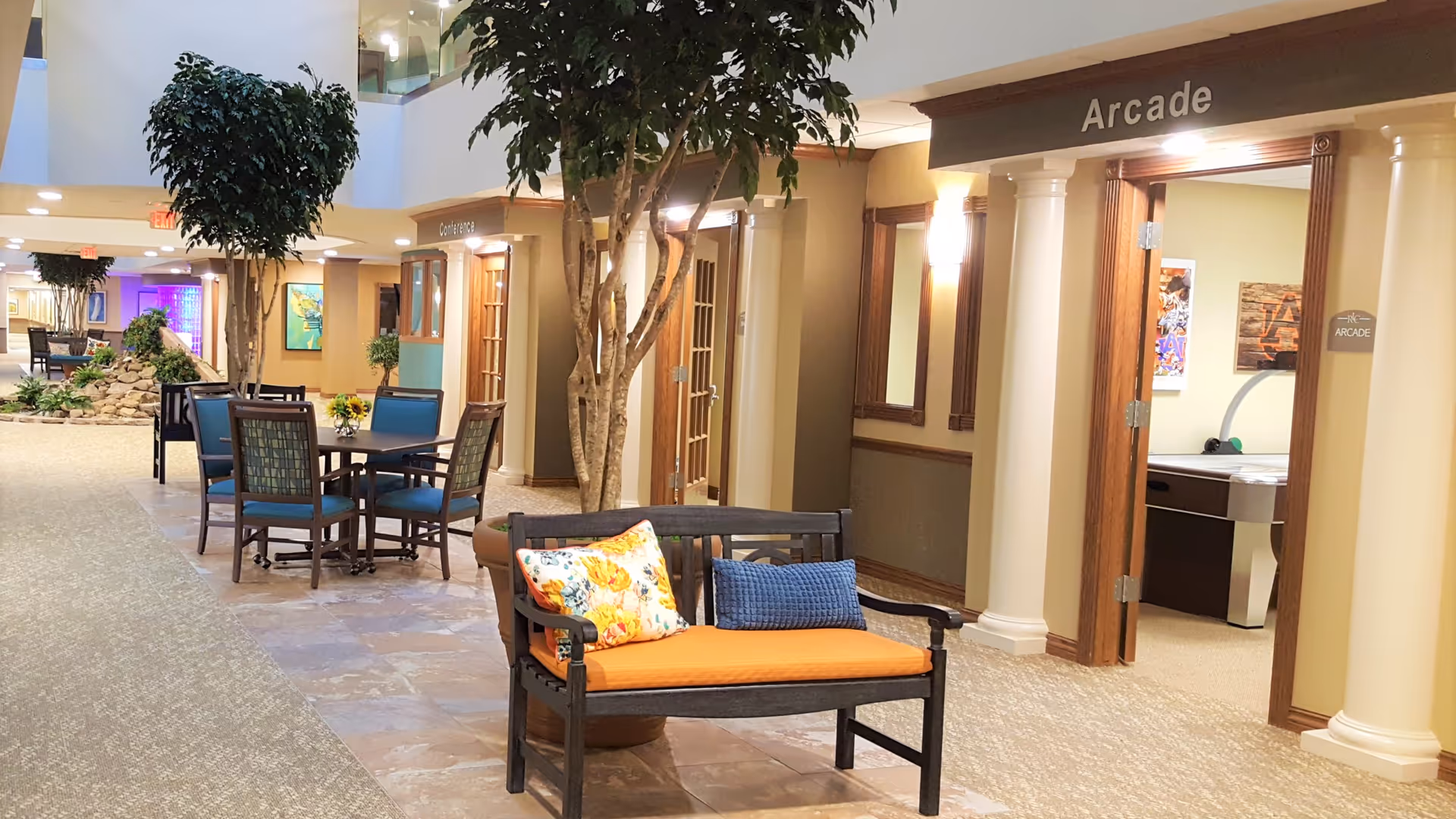 Interior hallway of a retirement community with seating areas, potted trees, and doors labeled Conference and Arcade. The Arcade room contains a game table visible through the open door.