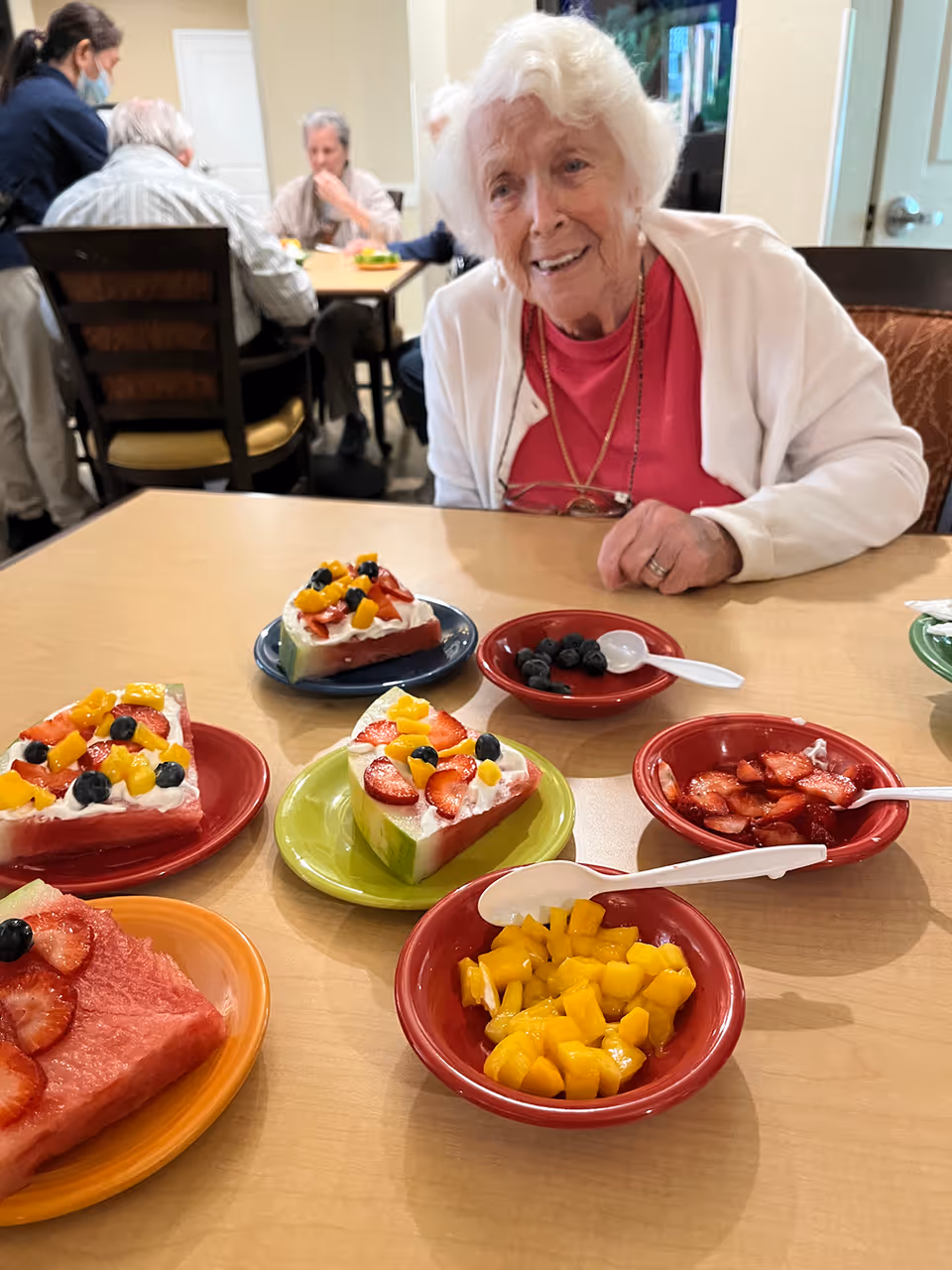 An elderly woman with white hair and a white cardigan sits at a table with several plates of fruit-topped desserts and bowls of fresh fruit in front of her. In the background, other elderly people are seated at tables, some engaged in conversation, in a communal dining area.