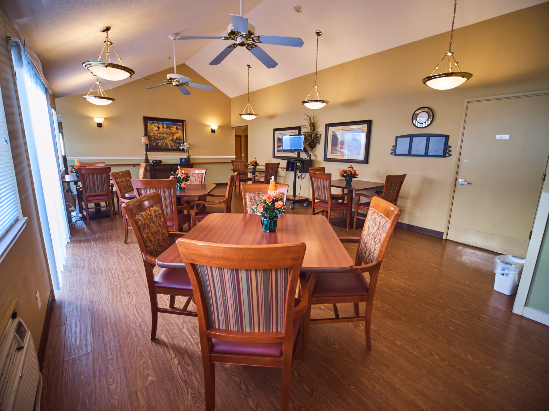 A dining room in a senior living facility with several wooden tables and chairs arranged neatly. Each table has a small floral centerpiece. The room has wood flooring, beige walls, ceiling fans, and hanging light fixtures. There are framed pictures on the walls and a computer workstation in the background.
