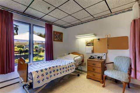 Hospital-style single bed in a furnished resident room with window and pink curtains.
