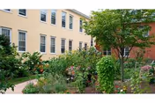 Landscaped courtyard garden with pathways, flowers, and trees beside a light-colored multi-story building.