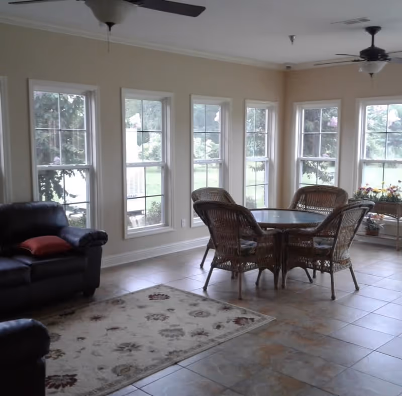 A bright room with multiple tall windows showing greenery outside. The room features a glass-top round table surrounded by four wicker chairs with cushions. There is a black leather sofa with a red pillow on the left side and a floral patterned rug on the tiled floor. Ceiling fans with lights hang from the ceiling, and a small table with flowers is placed near the windows.