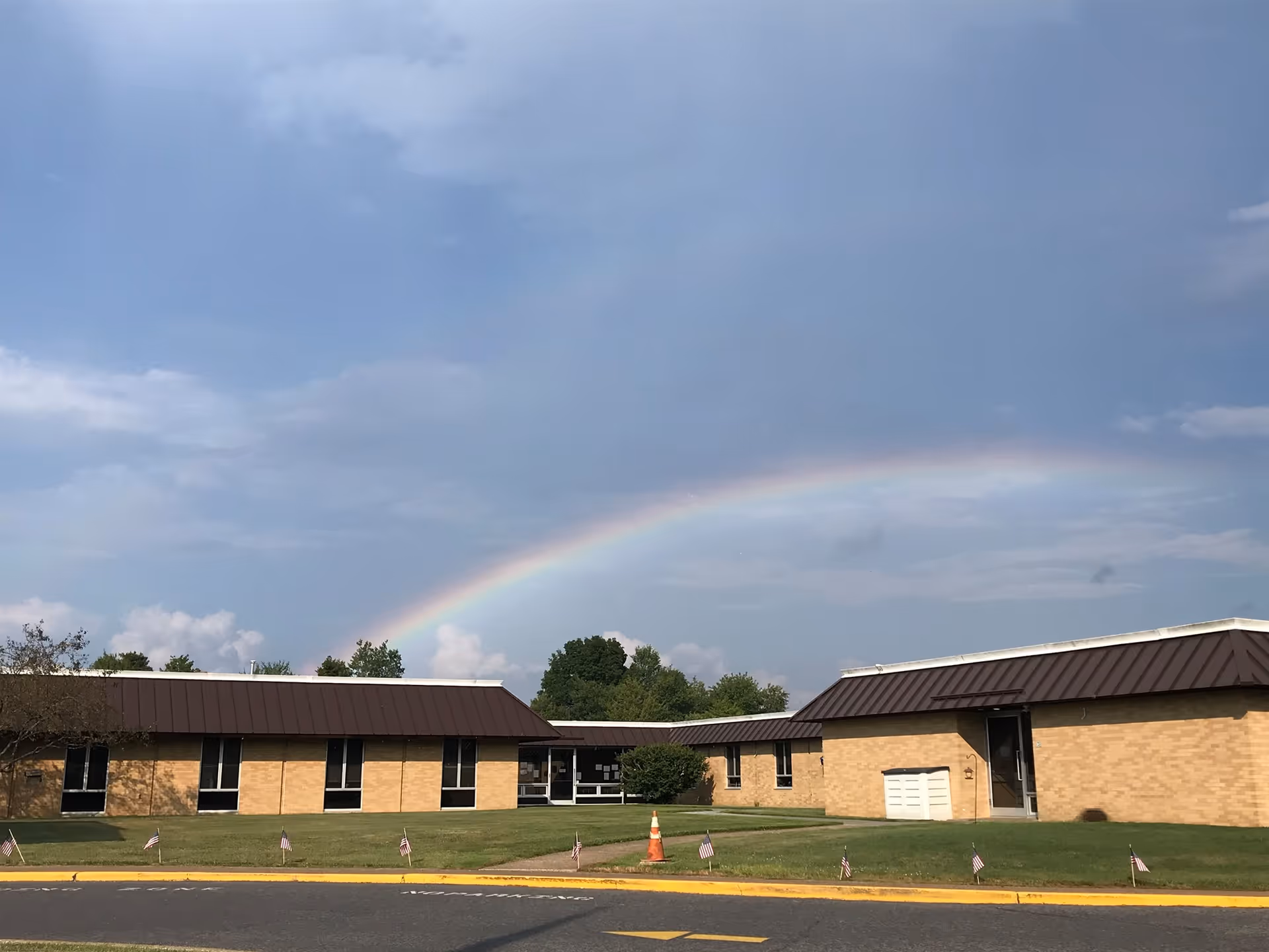 Exterior view of a single-story brick building with a brown roof under a partly cloudy sky with a visible rainbow. Small American flags line the grass near the sidewalk in front of the building.