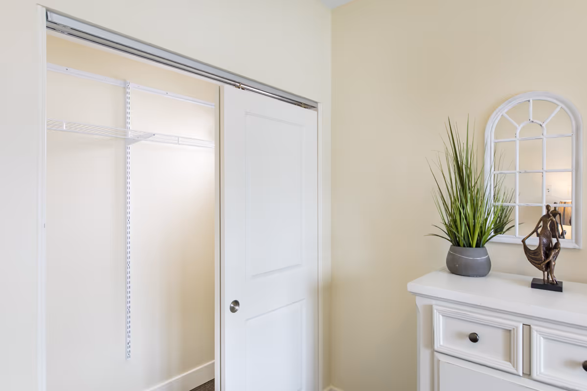 A corner of a room with a white sliding closet door partially open, revealing an empty closet with a wire shelf. Next to the closet is a white dresser with drawers, topped with a potted green plant and a small decorative sculpture. Above the dresser is a white-framed mirror with an arched windowpane design. The walls are painted a light beige color.