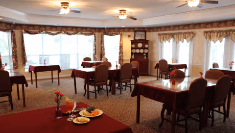 A dining room with multiple tables covered in red tablecloths, each set with chairs. One table in the foreground has a plate of food, a glass of orange juice, and a small vase with flowers. The room has large windows with curtains, ceiling fans with lights, and a cabinet with decorative plates in the background.
