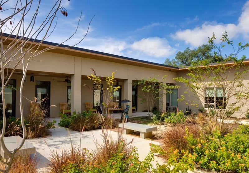 Outdoor courtyard area at The Watermark at Southpark Meadows featuring a paved walkway, benches, various plants and small trees, and a building with a covered patio and rocking chairs under a partly cloudy sky.