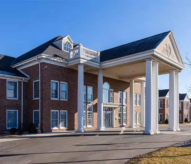 Exterior view of a two-story brick building with white columns supporting a large covered entrance, clear blue sky in the background.