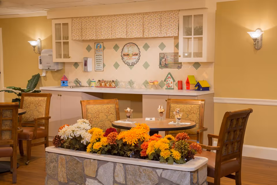 A cozy dining area with round tables and upholstered chairs, a large planter of colorful flowers in the foreground, and a small kitchenette with cabinets in the background.