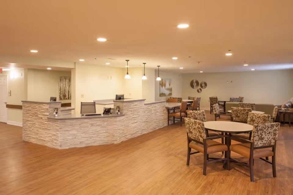 Interior view of a senior living facility common area featuring a reception desk with stone facade and multiple pendant lights above. The room has several round tables with cushioned chairs arranged around them, wooden flooring, and neutral-colored walls decorated with artwork and wall hangings.