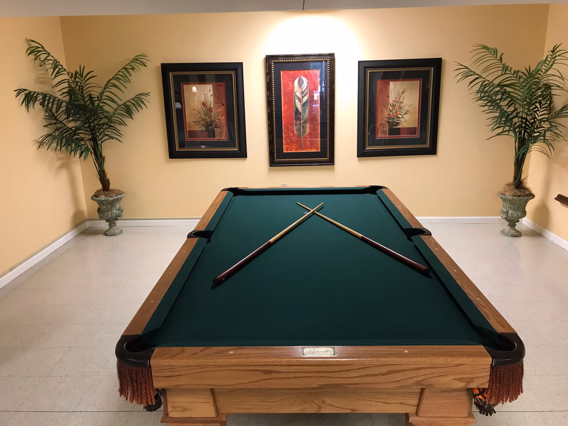 A pool table with green felt and two crossed pool cues resting on it, set in a room with beige walls. The wall behind the pool table features three framed artworks, with the center one depicting a large feather and the two side ones showing floral arrangements. There are two large potted plants placed symmetrically in the corners of the room.