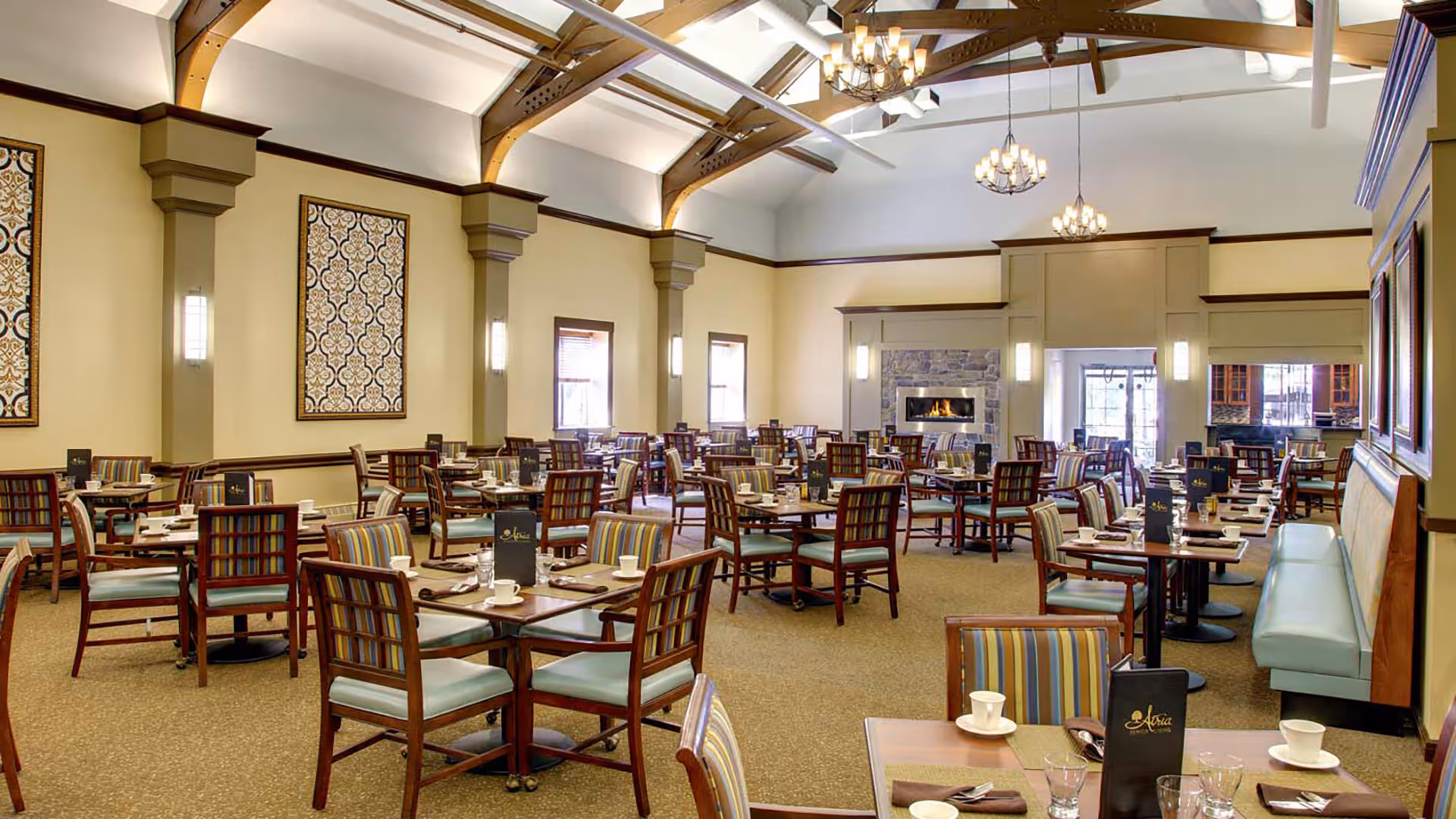 A spacious dining room with multiple wooden tables and chairs arranged neatly. Each table is set with cups, glasses, and menus. The room features high ceilings with exposed wooden beams, chandeliers, wall sconces, and large decorative wall panels. There is a stone fireplace at the far end of the room and a seating bench along one wall.