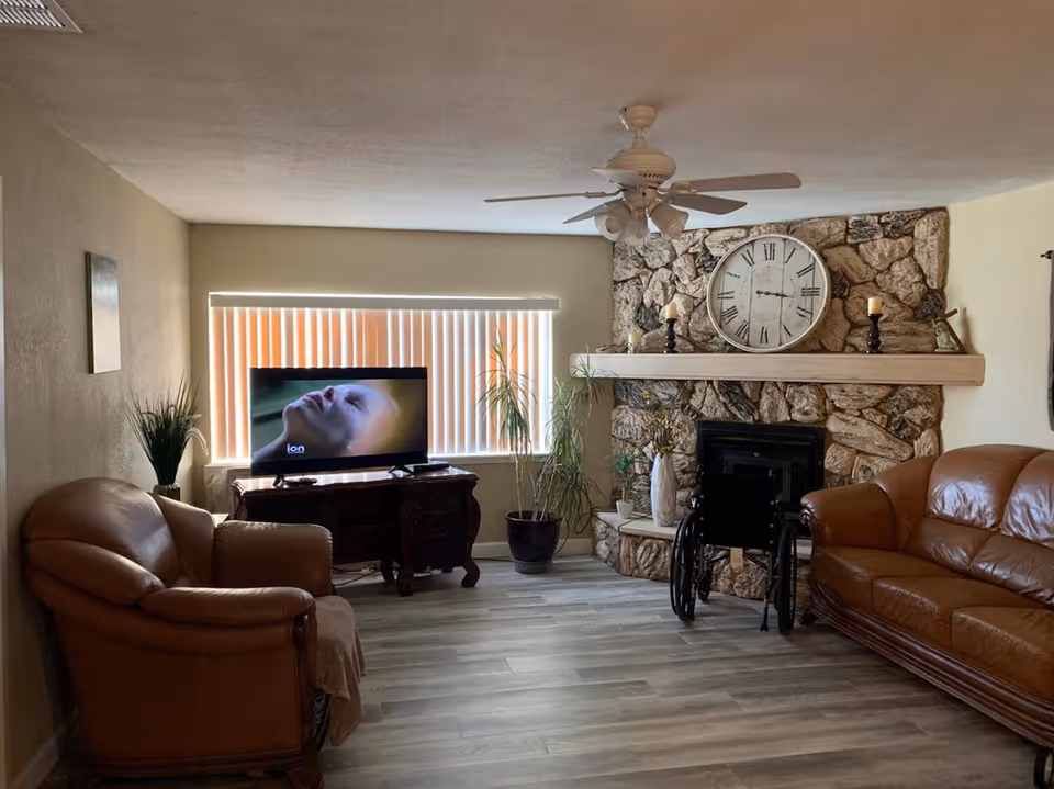 A cozy living room with two brown leather sofas, a wooden TV stand with a television showing a close-up of a person's face, a stone fireplace with a large clock and candles on the mantel, a ceiling fan with lights, and several potted plants near a window with vertical blinds.