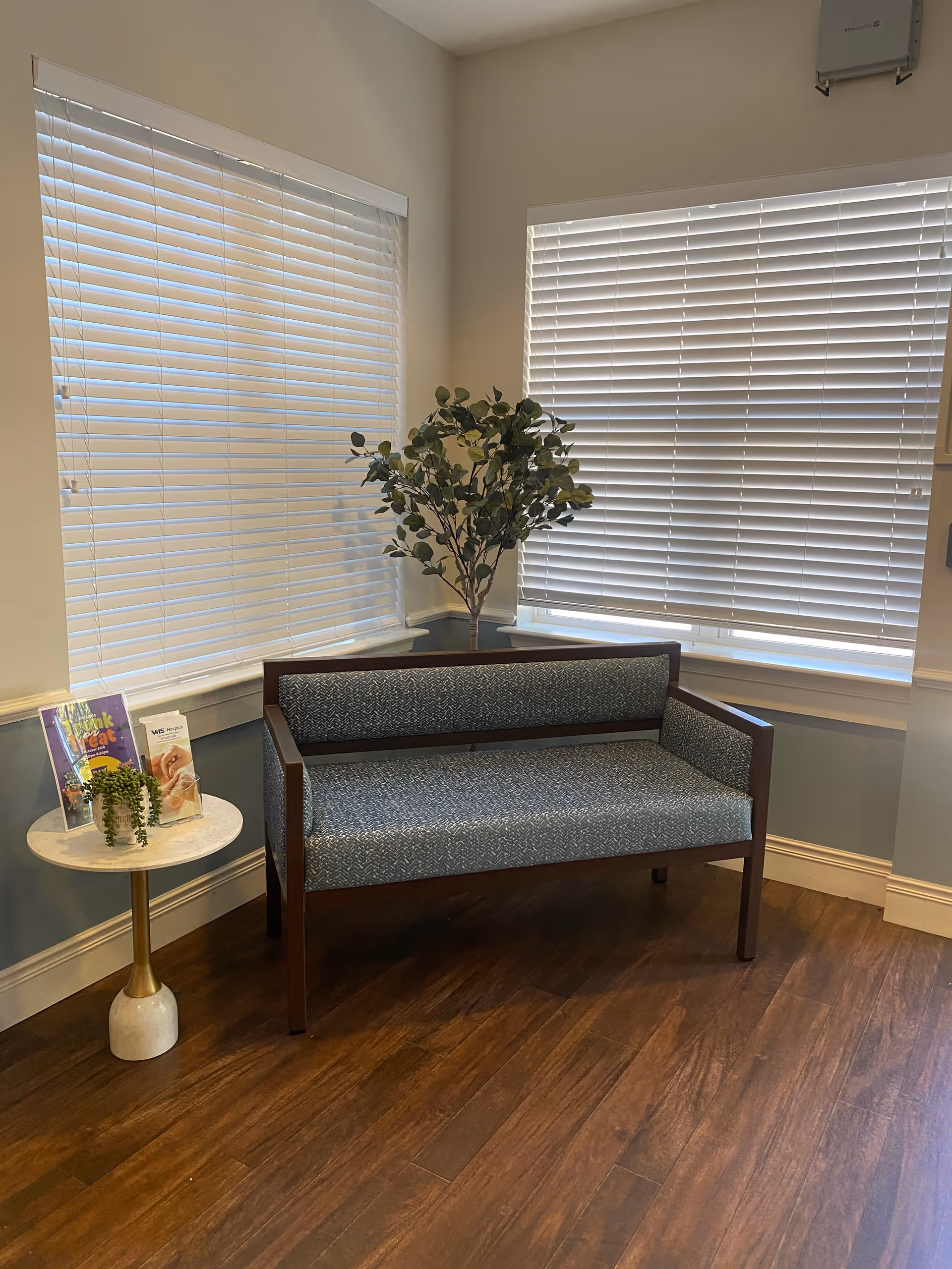 A small seating area with a blue patterned bench against a corner with two windows covered by white blinds. Next to the bench is a small round side table with a marble top and gold base, holding a small potted plant and some brochures. A tall green leafy plant is placed in the corner behind the bench. The floor is wooden and the walls are painted light blue and white.