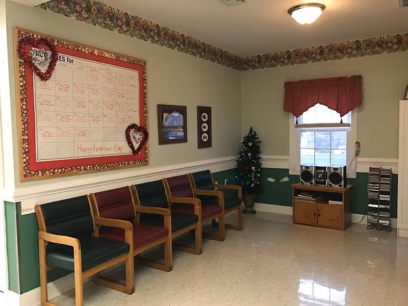 A small assisted-living common room with a row of wooden-framed chairs, a bulletin board, a small decorated Christmas tree, and a stereo on a cabinet by a window.