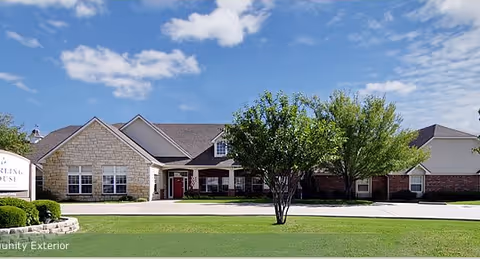 Exterior view of a single-story senior living facility building with a stone and brick facade, a well-maintained lawn, and a few trees under a blue sky with some clouds.