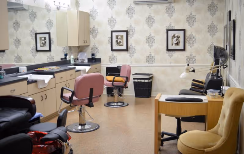 Interior of a salon area with two pink salon chairs in front of a counter with sinks and mirrors. The room has beige patterned wallpaper, framed pictures on the wall, a black laundry basket, a wooden manicure table with a yellow chair, and a black hair dryer chair.