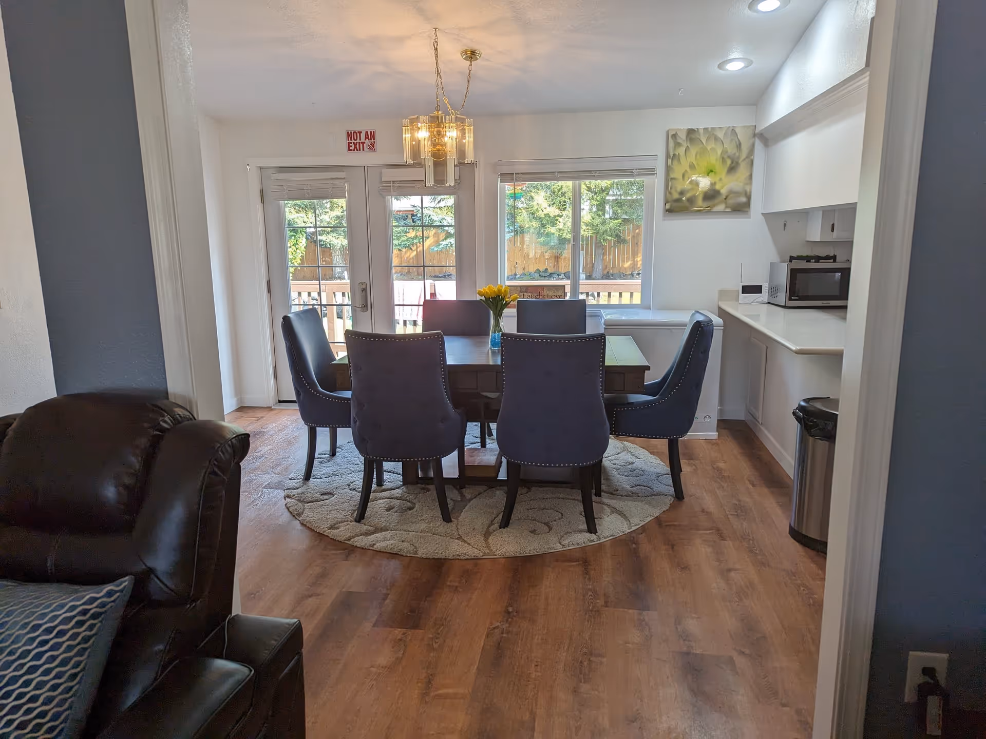 Dining area with a wooden table surrounded by six blue upholstered chairs on a round patterned rug. A vase with yellow flowers is on the table. Behind the table, there are glass doors and a window showing a fenced backyard with trees. To the right, there is a kitchen counter with a microwave and a trash can underneath. A floral wall art hangs above the counter.